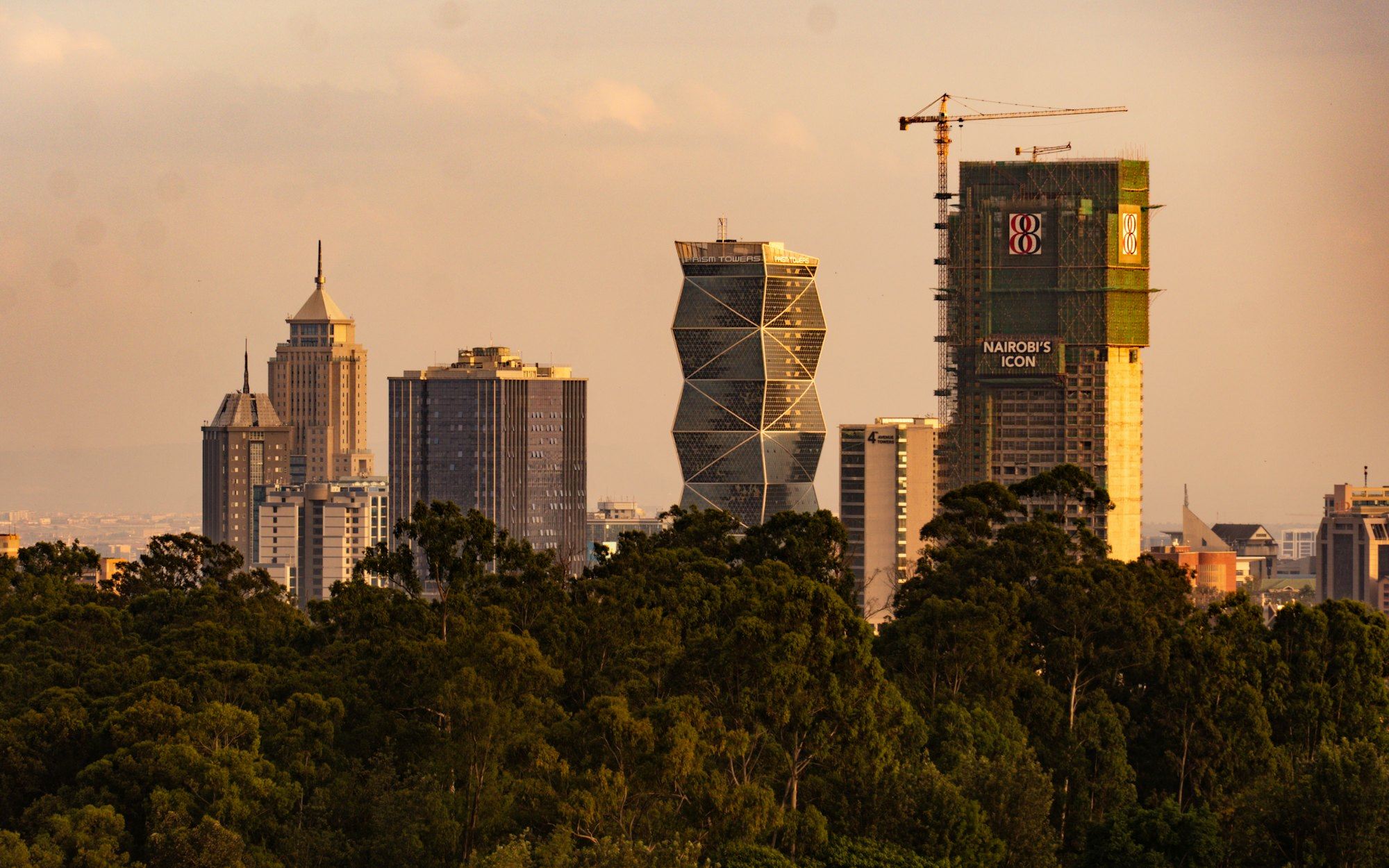 The Nairobi skyline at dusk, with completed towers and a high-rise under construction rising above the tree line.